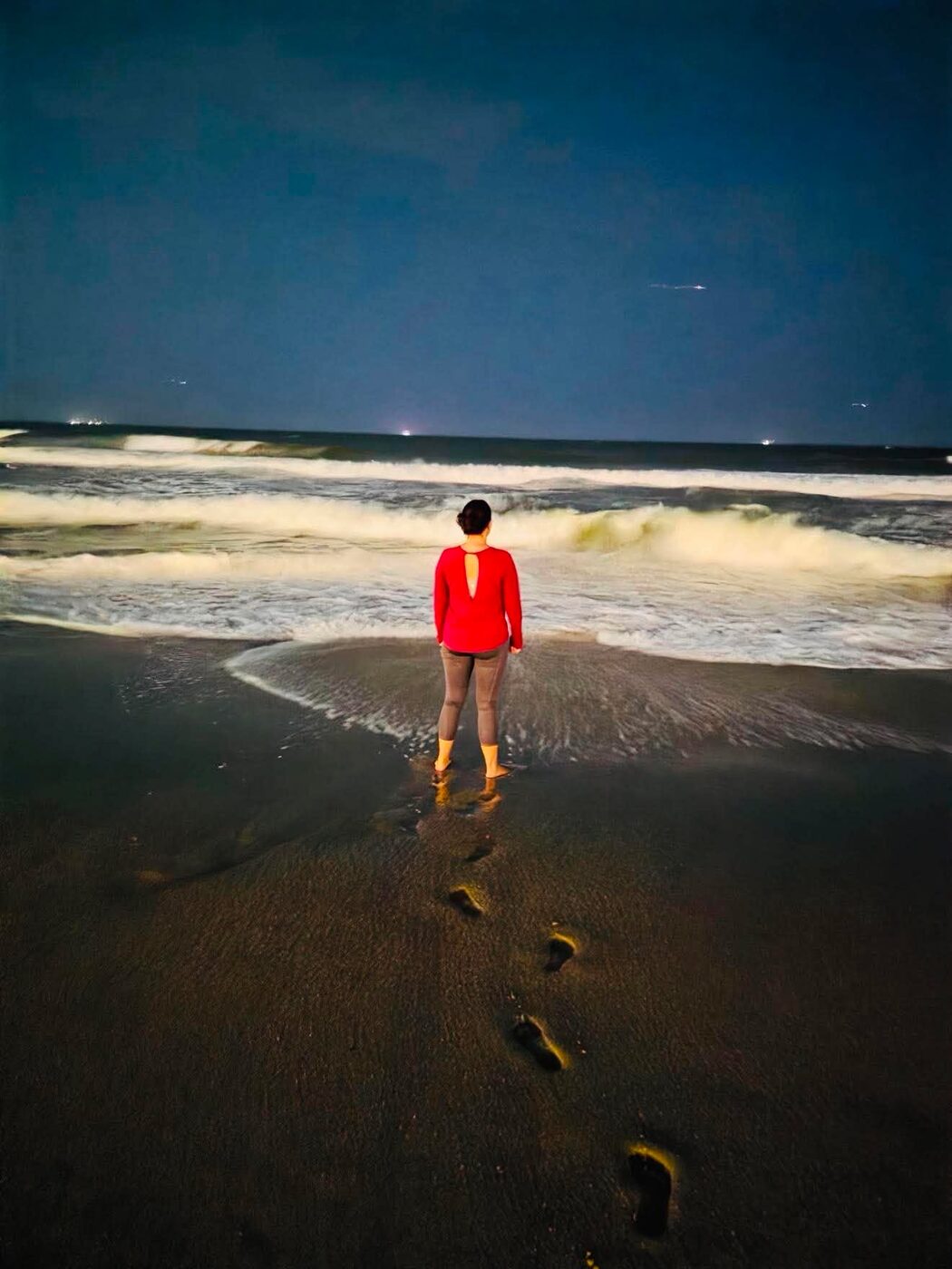 A person standing at the ocean's edge at night, footprints in the sand behind them